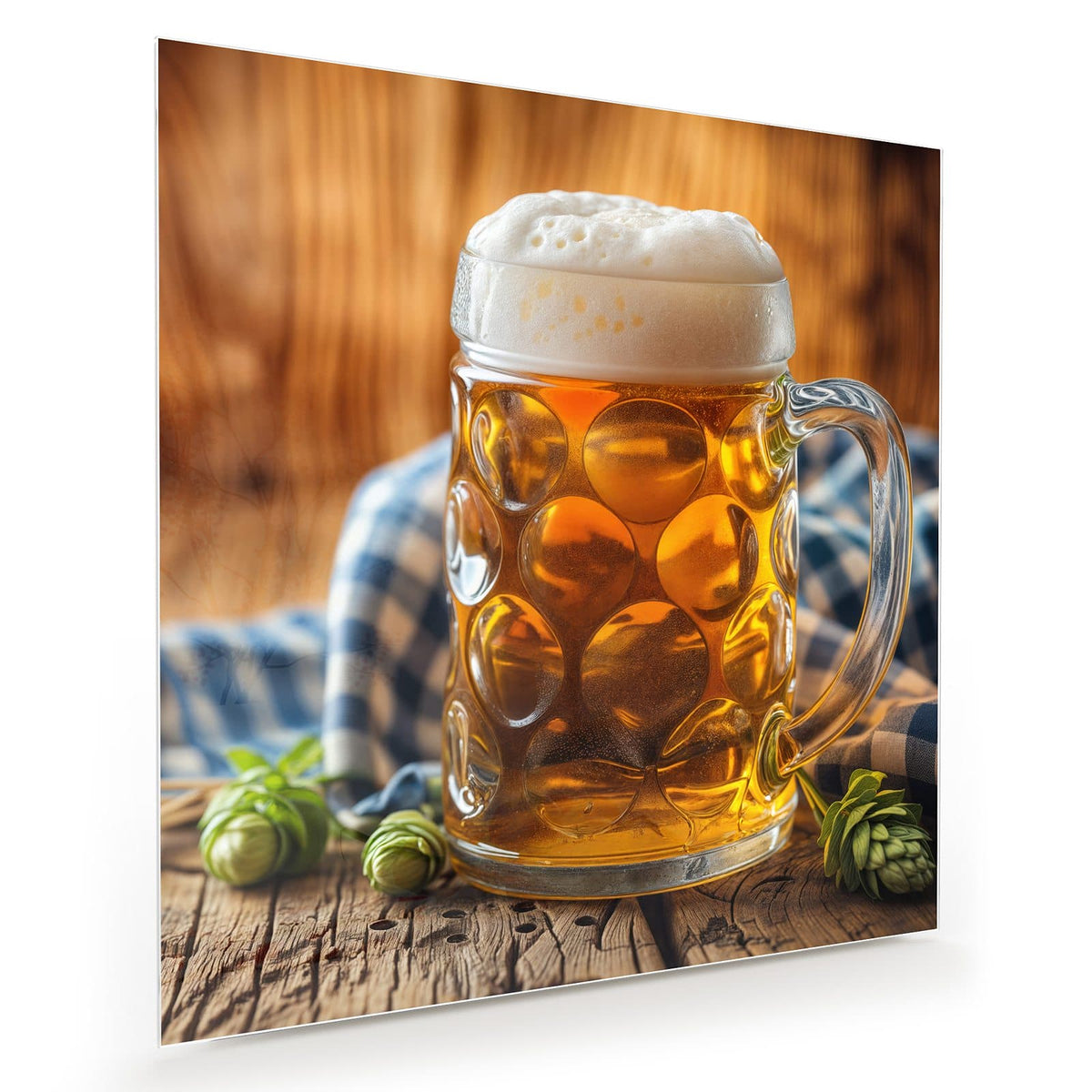Gedellter Glaskrug mit goldenem Bier und hoher Schaumkrone, davor Hopfendolden, dahinter ein blau-weiß kariertes Tuch.