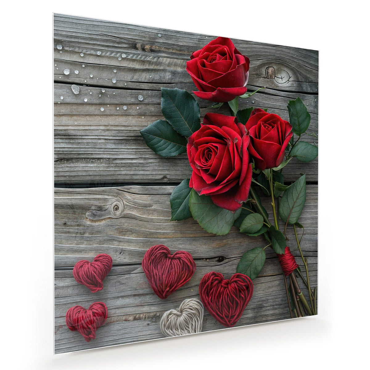 Drei tiefrote Rosen mit Blättern auf grauen Holzdielen, darunter vier aus rotem und beigem Garn gewickelte Herzen