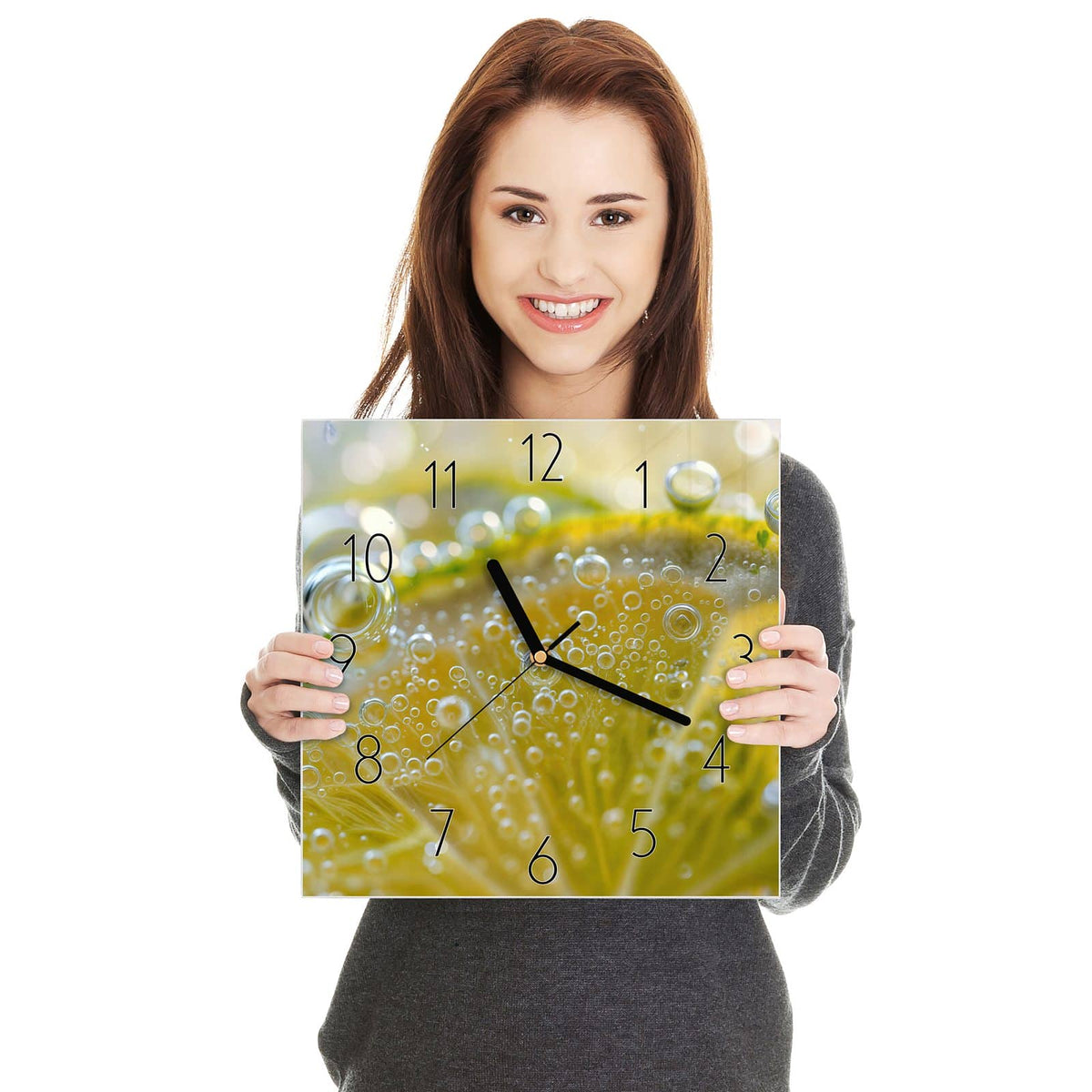 Quadratische Wanduhr aus Glas Zitrone im Sprudel in zwei Händen gehalten – Größenvergleich, 30 × 30 cm