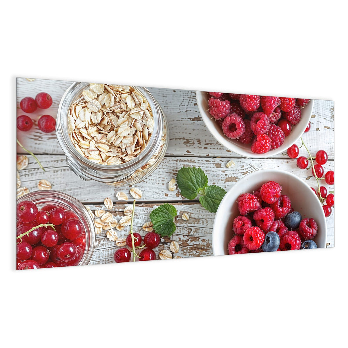 Zwei weiße Schalen mit Himbeeren, Blaubeeren und Johannisbeeren, dazu Haferflocken und Minze auf weißem Holz